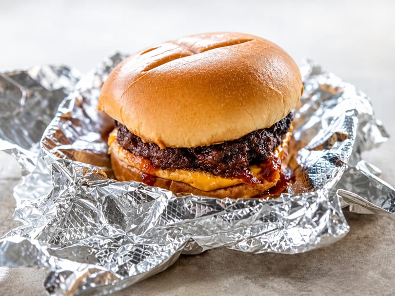 Juicy cheeseburger with melted cheese and sauce, wrapped in crinkled aluminum foil on a gray surface.