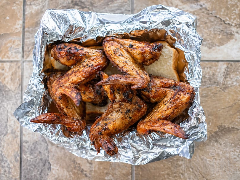 Grilled chicken wings with crispy skin and seasoning, served in a foil-lined container with bread.